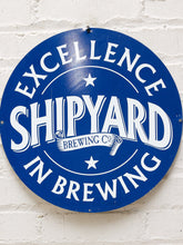 Load image into Gallery viewer, A round vintage-style blue and white metal sign reading 'Excellence Shipyard Brewing Co. in Brewing' mounted on a white painted brick wall.
