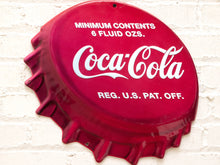 Load image into Gallery viewer, A red bottle-cap-shaped Coca-Cola sign with white lettering, mounted against a white painted brick wall.
