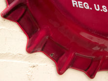 Load image into Gallery viewer, A close-up of a glossy deep red bottle-cap-shaped wall sign mounted on a white brick wall, with partial white text reading 'REG. U.
