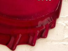 Load image into Gallery viewer, A close-up of a glossy deep red bottle-cap-shaped wall sign or decorative plaque against a white textured wall.
