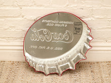 Load image into Gallery viewer, A vintage-style metal Coca-Cola bottle-cap shaped sign or tray with a distressed silver finish, red rim, and embossed Coca-Cola script, leaning against a white brick wall on a woven rug.
