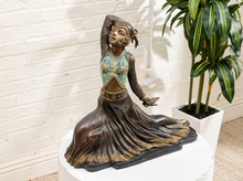 Load image into Gallery viewer, A bronze-finished statue of a dancing woman in a flowing dress with turquoise and gold painted accents, displayed on a white pedestal in front of a white brick wall next to a potted plant.
