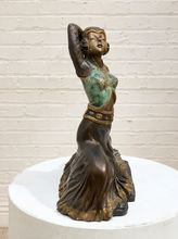 Load image into Gallery viewer, A bronze-finished sculpture of a dancing woman with a turquoise and gold painted bodice, displayed on a white round pedestal against a white brick wall.
