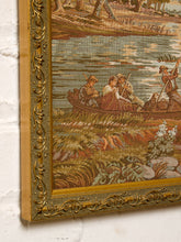Load image into Gallery viewer, Close-up of an ornate framed textile wall hanging or tapestry showing a historical river scene with figures in a boat, mounted against a white wall.
