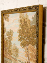 Load image into Gallery viewer, A framed landscape tapestry featuring trees and a soft sky, shown at an angle against a white brick wall. The tapestry has a warm beige and brown color palette, and it is enclosed in an ornate gold-colored frame.
