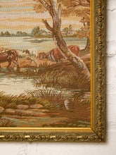 Load image into Gallery viewer, A close-up of an ornate framed tapestry or wall hanging showing a pastoral landscape with cows near water, mounted against a white textured wall.
