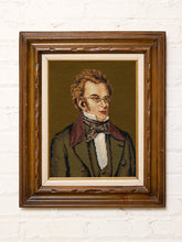 Load image into Gallery viewer, Vintage framed needlepoint portrait depicting a 19th-century gentleman with curly brown hair, round spectacles, a dark suit, and a patterned bow tie against a deep brown background, displayed in an ornate dark wood frame.
