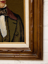 Load image into Gallery viewer, Close-up of a framed needlepoint portrait showing part of a person in a brown coat and white shirt, with a thick carved dark wood frame and cream mat against a white brick wall.
