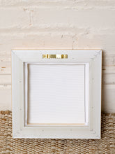 Load image into Gallery viewer, A square white wooden picture frame with a gold metal hanger at the top, shown against a white painted brick wall and a woven jute rug.
