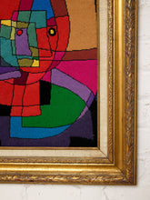 Load image into Gallery viewer, A close-up of a framed abstract needlepoint or textile artwork with bold geometric shapes in red, green, blue, purple, pink, yellow, and black, set in an ornate gold frame against a white brick wall.
