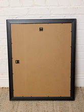 Load image into Gallery viewer, The back of a black picture frame leaning against a white painted brick wall on a woven rug. The backplate is tan cardboard with pre-drilled holes and small black mounting tabs.
