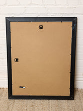 Load image into Gallery viewer, The back of a black picture frame with a brown cardboard backing, leaning against a white painted brick wall on a woven jute rug.
