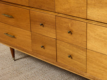 Load image into Gallery viewer, Close-up of a mid-century modern wooden dresser with multiple drawers, brass pulls, and a tapered leg on a textured carpet.
