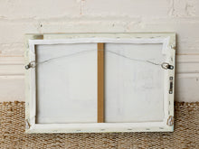 Load image into Gallery viewer, The back side of a rectangular framed canvas print, showing the wire hanging system, metal clips, and wooden stretcher bars, set against a white brick wall and woven rug.
