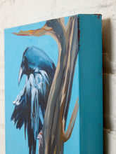 Load image into Gallery viewer, A close-up of a canvas painting featuring a raven perched on a tree trunk against a bright blue background, shown leaning against a white brick wall.

