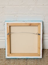 Load image into Gallery viewer, The back of a square canvas print showing the wooden frame, canvas backing, and hanging wire hardware, set against a white brick wall and woven rug.

