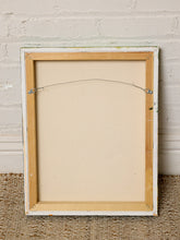 Load image into Gallery viewer, The back of a square framed art print or canvas, showing a light wood frame with white paint, a wire hanger, and a beige backing board, set against a white brick wall and woven rug.
