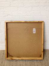 Load image into Gallery viewer, The back of a square wooden picture frame with a light natural wood border and a brown cardboard backing, standing on a woven rug against a white painted brick wall.
