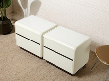 Load image into Gallery viewer, Two modern cream-colored leather storage ottomans with two drawers each, positioned side-by-side on a textured beige carpet against a white brick wall.
