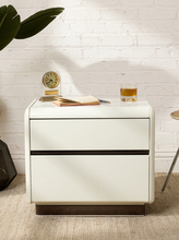 Load image into Gallery viewer, A modern two-drawer nightstand in glossy white with a dark wood base, styled in a room setting with a white brick wall, a plant leaf, a small clock, an open notebook, and a glass of whiskey.
