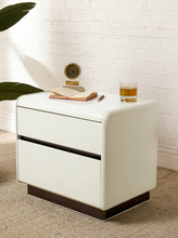Load image into Gallery viewer, A modern two-drawer nightstand in off-white faux leather with dark brown base and drawer accents, styled with an open book, pen, small clock, and a glass of iced tea on top against a white brick wall.
