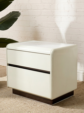 Load image into Gallery viewer, A modern two-drawer nightstand with a glossy off-white finish, dark brown base, and a thin dark horizontal accent line between the drawers, set against a white brick wall and beige carpet.
