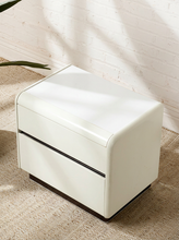 Load image into Gallery viewer, A modern cream-colored two-drawer nightstand with a glossy finish, placed on a textured beige rug against a white brick wall with soft leaf shadows.

