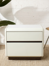 Load image into Gallery viewer, A modern two-drawer nightstand in a warm off-white finish with a dark horizontal accent strip between the drawers and dark wood feet, set against a white brick wall with a plant leaf in the foreground.

