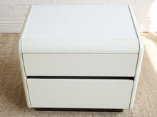Load image into Gallery viewer, A small white two-drawer nightstand or storage cabinet with rounded edges, shown on a woven rug against a white brick wall.
