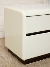 Load image into Gallery viewer, A close-up of a cream-colored two-drawer nightstand or cabinet with dark brown base trim, set on a textured beige carpet against a white brick wall.
