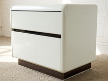 Load image into Gallery viewer, A two-drawer nightstand with a glossy white finish, dark brown base, and black drawer pulls, set on a textured beige carpet against a white brick wall.
