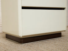 Load image into Gallery viewer, Close-up of the lower portion of a cream-colored two-drawer cabinet or nightstand with a dark brown wooden base, resting on a textured beige carpet.
