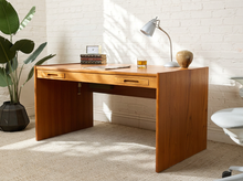 Load image into Gallery viewer, Image 2 of 22: A mid-century modern wooden writing desk with two drawers, styled in a bright room with a white brick wall, a potted plant, and a silver desk lamp.
