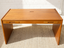 Load image into Gallery viewer, Image 8 of 22: A mid-century modern wooden writing desk with a warm honey-toned finish, featuring two pull-out drawer fronts and a small center slot on the tabletop, set on a woven rug against a white brick wall.
