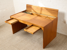Load image into Gallery viewer, Image 17 of 22: A mid-century modern wooden writing desk with a warm honey finish, featuring a lift-up back panel, a top tray, and two pull-out side drawers, set against a white brick wall and beige carpet.

