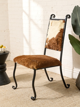 Load image into Gallery viewer, Image 2 of 15: A rustic accent chair with a black wrought-iron frame, featuring a brown faux-fur seat cushion and a cowhide-patterned backrest, set against a white brick wall.
