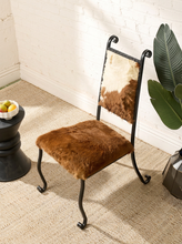 Load image into Gallery viewer, Image 3 of 15: A rustic accent chair with a brown and white cowhide seat and backrest, supported by a black metal frame, set on a beige carpet next to a white brick wall.
