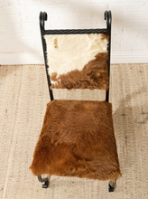 Load image into Gallery viewer, Image 4 of 15: A rustic chair with a black metal frame and cowhide upholstery, featuring a brown and cream hide backrest and a solid medium-brown hide seat, set on a woven jute rug against a white brick wall.
