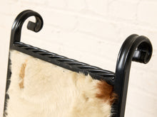 Load image into Gallery viewer, Image 8 of 15: Close-up of a black metal chair frame with a curved scrollback design and a cream-colored cowhide seat, set against a white brick wall background.
