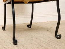 Load image into Gallery viewer, Image 13 of 15: Close-up of a black metal stool or bench with curved legs and a tan upholstered seat, shown on a beige carpet against a white wall.
