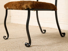 Load image into Gallery viewer, Image 14 of 15: A rustic stool with a brown faux-fur or sheepskin seat cushion and black metal scroll legs, shown on a beige carpet against a white brick wall.
