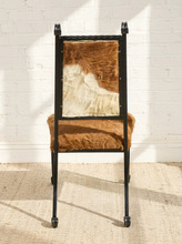 Load image into Gallery viewer, Image 6 of 15: A black metal chair with a cowhide upholstered seat and backrest, set against a white brick wall on a woven rug.
