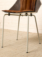 Load image into Gallery viewer, Image 15 of 16: A mid-century modern style chair with a brown leather seat and backrest, supported by a light gray metal frame, set on a textured beige rug against a white brick wall.
