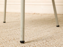 Load image into Gallery viewer, Image 16 of 16: Close-up of a light gray metal table leg with a black rubber cap on a beige woven rug, with other legs blurred in the background.
