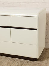 Load image into Gallery viewer, Image 6 of 18: A close-up of a glossy off-white dresser with dark brown trim accents, set against a white brick wall and textured carpet.
