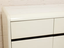 Load image into Gallery viewer, Image 9 of 18: Close-up of a glossy off-white cabinet or dresser showing part of the top surface, front drawer, and a thin black horizontal accent line, set against a white brick wall.
