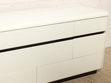Load image into Gallery viewer, Image 10 of 18: A close-up of a modern white dresser with six drawers and slim black horizontal handles, set against a white brick wall and carpeted floor.
