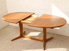 Load image into Gallery viewer, Image 6 of 28: A mid-century modern wooden extendable dining table with a warm medium-brown finish, shown partially open on a textured beige rug against a white brick wall.
