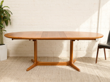 Load image into Gallery viewer, Image 7 of 28: A mid-century modern oval wooden dining table with a warm medium-brown finish, set against a white brick wall on a textured beige rug.
