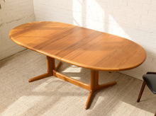 Load image into Gallery viewer, Image 8 of 28: An oval wooden dining table with a warm medium-brown finish and a pedestal base, shown in a room with a white brick wall and light-colored carpet.
